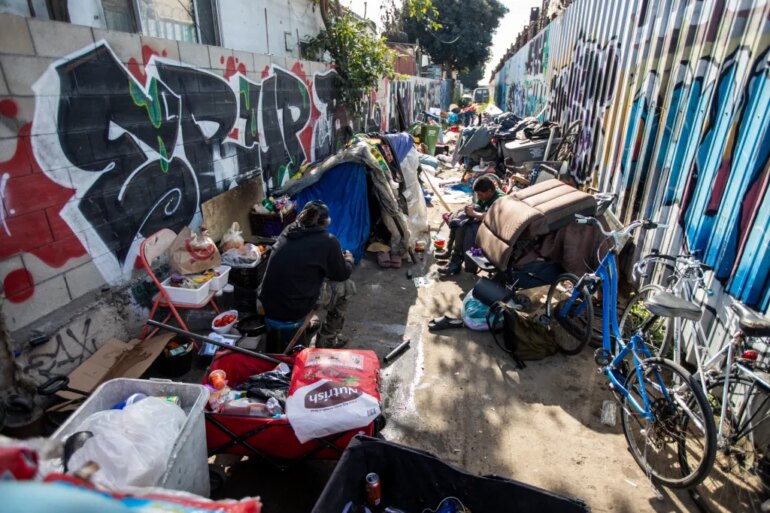 People sit among their belongings at an encampment in an alley.