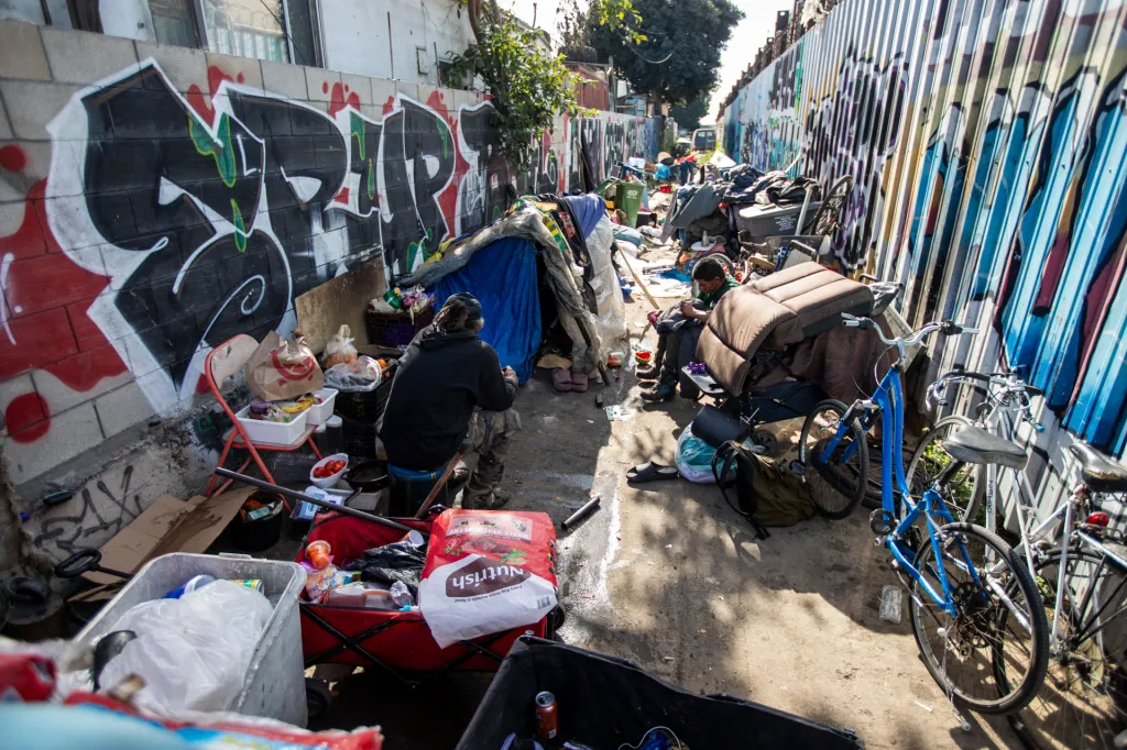 People sit among their belongings at an encampment in an alley.