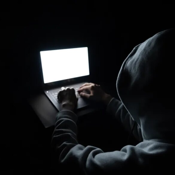 A hooded figure typing on a laptop with a bright white screen in a dark room.