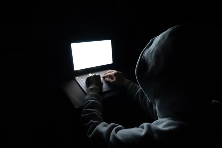 A hooded figure typing on a laptop with a bright white screen in a dark room.