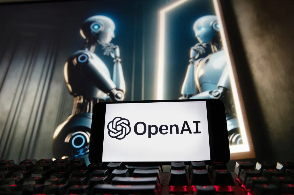 A cellphone displaying the OpenAI logo rests on a keyboard, with two robot figures on a computer monitor in the background.