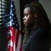 Portrait of a stressed young teenage girl looking out a window with an American flag in the background.