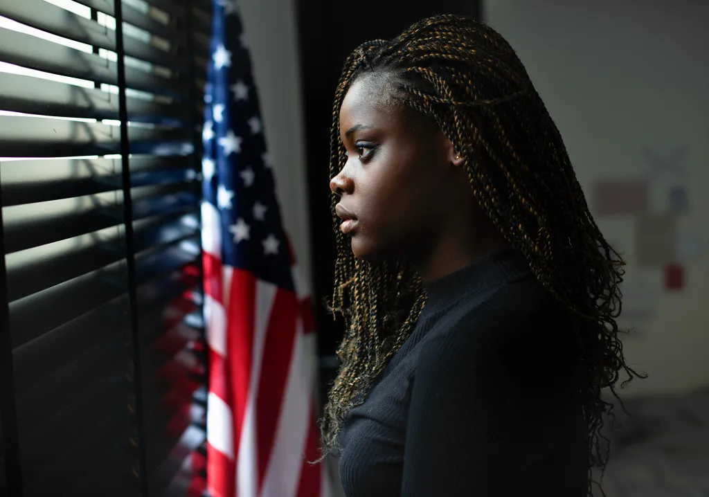 Portrait of a stressed young teenage girl looking out a window with an American flag in the background.